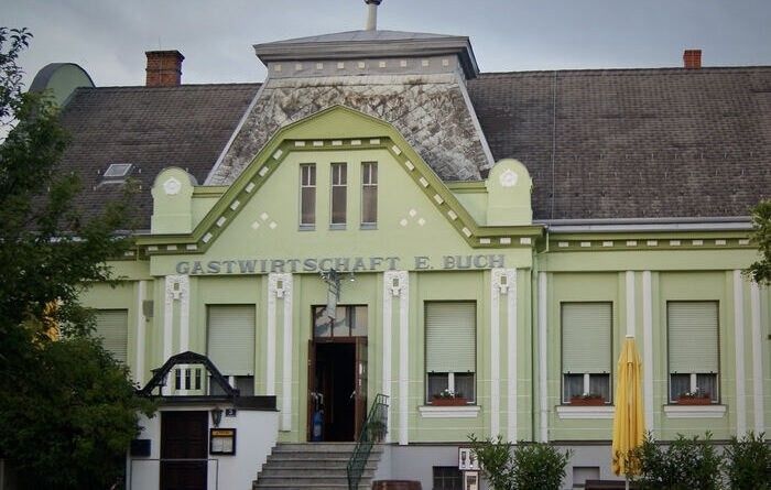 Ein grünes Gebäude mit weißen Verzierungen trägt eine Tafel mit der Aufschrift Gastwirtschaft E. Buch. Das Gebäude hat Fenster mit Rollläden und einen gelben Regenschirm davor.