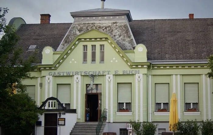 Ein grünes Gebäude mit weißen Verzierungen trägt eine Tafel mit der Aufschrift Gastwirtschaft E. Buch. Das Gebäude hat Fenster mit Rollläden und einen gelben Regenschirm davor.