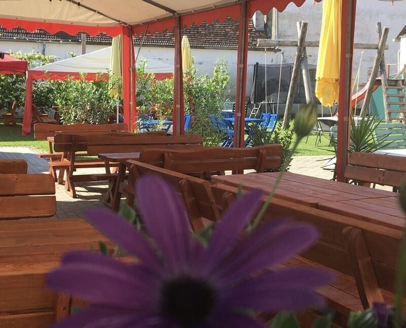 Ein Outdoor-Café mit Holztischen und Bänken unter einem Baldachin. Es gibt gelbe Regenschirme und Pflanzen. Einige Tische haben Blumen im Vordergrund.