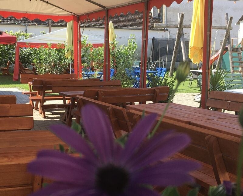 Ein Outdoor-Café mit Holztischen und Bänken unter einem Baldachin. Es gibt gelbe Regenschirme und Pflanzen. Einige Tische haben Blumen im Vordergrund.