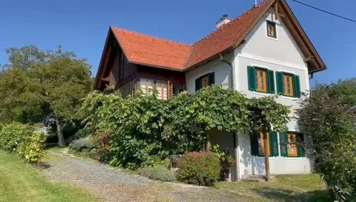 Ein weißes Haus mit rotem Dach, grünen Fensterläden und einer Einfahrt, umgeben von Grün und Pflanzen.
