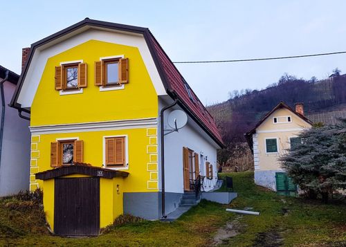 Ein gelbes Haus mit rotem Dach und braunen Fensterläden. Ein kleines Haus mit grünen Fensterläden steht in der Nähe.