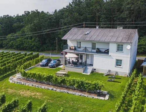 Eine Vogelperspektive eines weißen Hauses mit zwei Autos davor, einer Bank, einem Tisch mit einem Regenschirm und einer Terrasse mit Tisch und Stühlen sowie einer Satellitenschüssel.