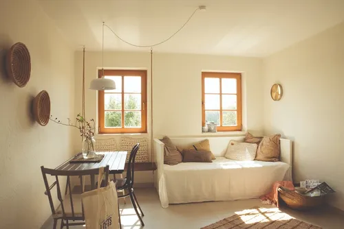 Ein gemütliches Zimmer mit weißen Wänden und Holzmöbeln. Ein Sofa mit Kissen steht in der Nähe des Fensters. Ein Tisch mit einer Tasche daneben und zwei Stühlen. Eine Pflanze in einer Vase steht auf dem Tisch.
