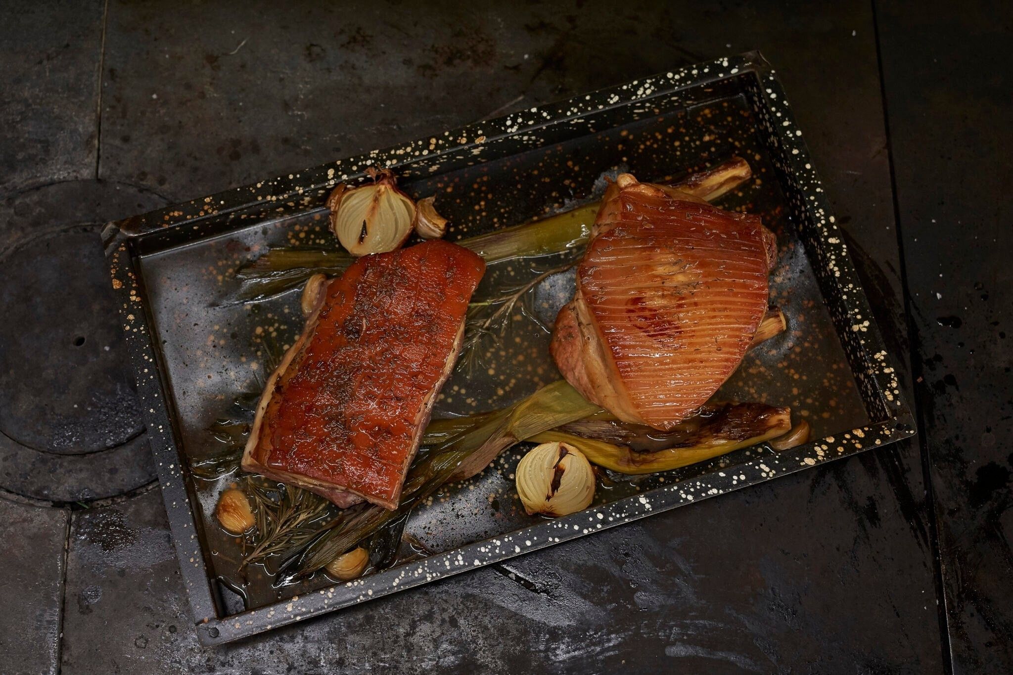 Ein schwarzes rechteckiges Tablett mit zwei glasierten Fleischstücken, Kräutern und Zwiebeln. Das Fleisch hat eine rötlich-braune Glasur und ist mit Kräutern garniert.