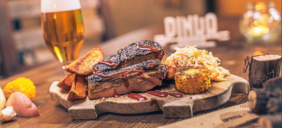 Ein Holzbrett mit Rippen, Süßkartoffel-Pommes, Maiskolben und Krautsalat, dazu ein Glas Bier. Dahinter steht ein großes Schild mit der Aufschrift 'PINO'.