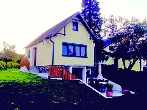 Ein Haus mit gelben Wänden, einem braunen Dach und blauen Fensterrahmen steht auf einem schiefen, grünen Hang, umgeben von Bäumen und Büschen.