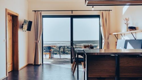 Ein moderner Essbereich mit einer großen Glastür, die zu einem Balkon führt, mit Tisch und Stühlen, einem montierten Fernseher und Blick auf den Ozean.
