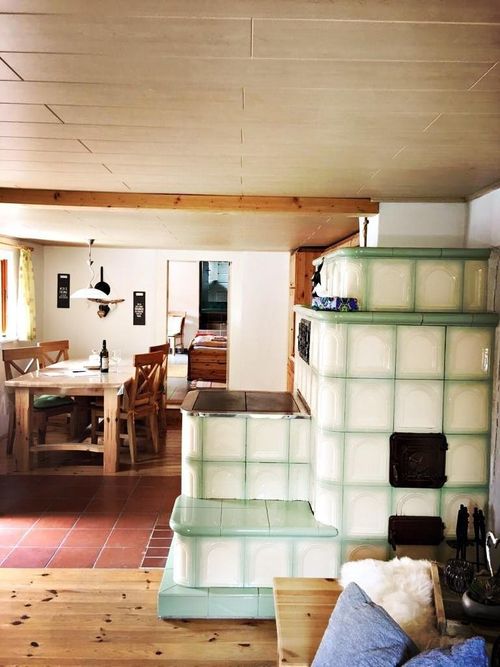 Ein gemütliches Esszimmer mit Holzmöbeln und gefliestem Boden. Ein Holztisch mit Flasche und Gläsern steht neben einer Holzwand mit einem großen Ofen. Eine Lampe hängt von der Decke, und ein Fenster mit gelben Vorhängen lässt natürliches Licht herein.
