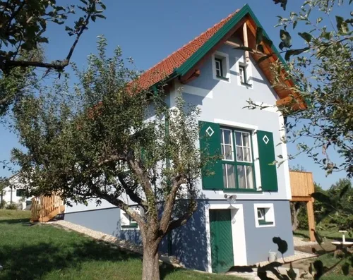 Ein Haus mit rotem Dach und grünen Fensterläden ist von einem Rasenbereich und Bäumen umgeben. Es verfügt über eine Holzterrasse, eine weiße Wand und einen kleinen Holzzaun.