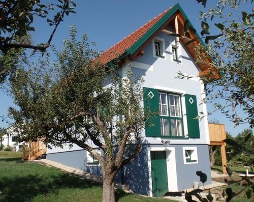 Ein Haus mit rotem Dach und grünen Fensterläden ist von einem Rasenbereich und Bäumen umgeben. Es verfügt über eine Holzterrasse, eine weiße Wand und einen kleinen Holzzaun.