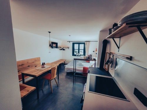 Ein Interieur eines kleinen Apartments mit einem Holztisch und Stühlen, einem Bett und einem Küchenbereich mit Waschbecken und Herd.