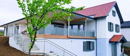 Ein modernes weißes Haus mit rotem Dach und hölzernen Balkongeländern, mit einem Baum und Treppen, die zum Eingang führen. Das Haus ist von einer Rasenfläche umgeben.