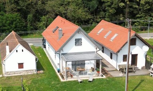 Eine Vogelperspektive von drei Häusern, eines mit einem roten Dach, ein anderes mit einem braunen Dach und ein drittes mit einer verglasten Terrasse, alle umgeben von grünem Gras und Bäumen.