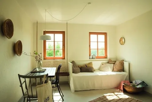 Ein gemütliches Zimmer mit einem weißen Sofa, Kissen und einem Tisch mit einer Pflanze in einer Vase. Es gibt zwei Fenster, einen Korb und dekorative Wandbehänge.