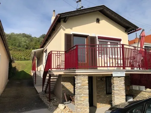 Ein beigefarbenes Haus mit rotem Balkon und Treppe, umgeben von Grün und einem Auto vor dem Haus.