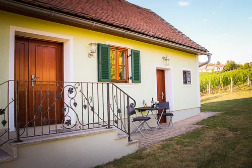 Ein gelbes Haus mit einem roten Dach und grünen Fensterläden. Ein Balkon mit schwarzem Geländer führt zu einem kleinen Essbereich im Freien.