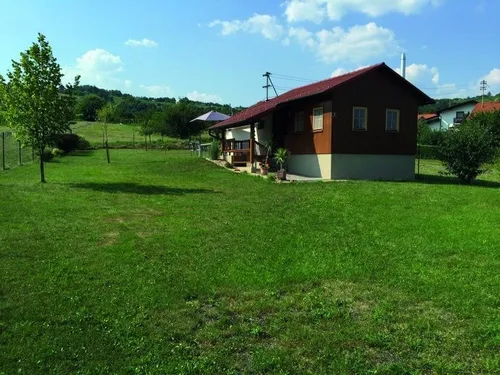 Ein kleines braunes Haus mit rotem Dach und weißer Umrandung steht auf einem üppigen grünen Rasen, mit einem kleinen Vordach und Topfpflanzen davor. Bäume und Sträucher umgeben das Haus, mit einem Feld und entfernten Häusern im Hintergrund.