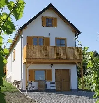 Ein zweistöckiges Haus mit einem Holzbalkon, gelben Fensterläden und einer Holztür. Zwei Stühle und ein Fass stehen vor dem Eingang.