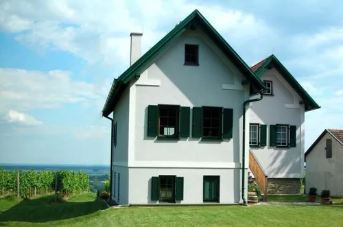 Ein weißes Haus mit grünen Fensterläden und einem grünen Dach steht auf einem grünen Hügel, mit einem Weinberg und einem kleineren Haus in der Nähe.