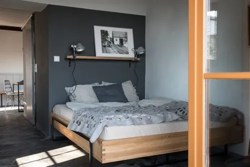 Ein Schlafzimmer mit einem Bett, Kissen und einer Decke. Ein Regal mit einem Bilderrahmen und Lampen ist an der Wand montiert. Ein Stuhl und ein Tisch stehen in der Ecke.