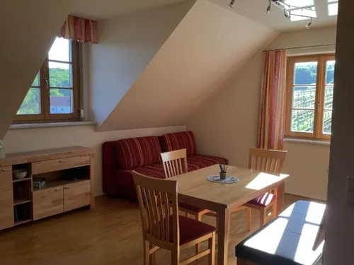 Ein Dachzimmer mit einem hölzernen Esstisch und Stühlen, einem roten Sofa und zwei Fenstern. Der Raum hat Holzböden und Wände.