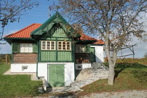 Ein rustikales Haus mit grünen Fensterläden und einem roten Dach steht in einer ländlichen Gegend, umgeben von Gras und einem Baum.