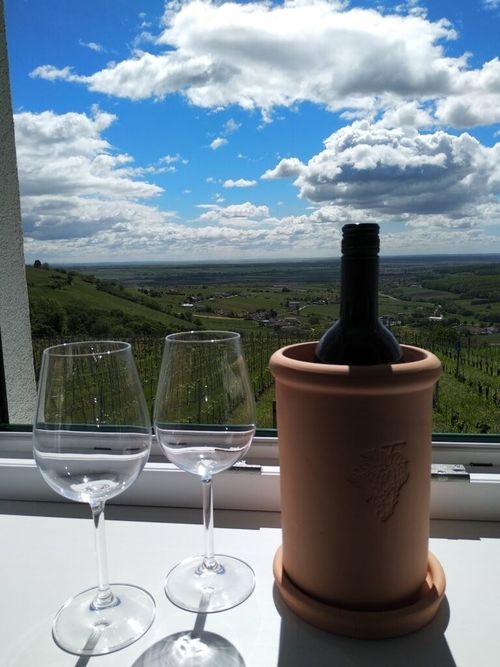 Zwei Weingläser stehen neben einer Flasche in einem Terrakotta-Flaschenkühler, gesehen aus einem Fenster mit einer wunderschönen Landschaft.