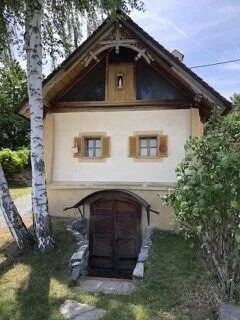 Ein kleines, traditionelles Haus mit einer Holztür und drei Fenstern, eingebettet zwischen Bäumen und Grün. Das Dach ist steil geneigt und die Wände sind in einer hellen Farbe gestrichen.
