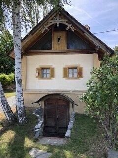 Ein kleines, traditionelles Haus mit einer Holztür und drei Fenstern, eingebettet zwischen Bäumen und Grün. Das Dach ist steil geneigt und die Wände sind in einer hellen Farbe gestrichen.