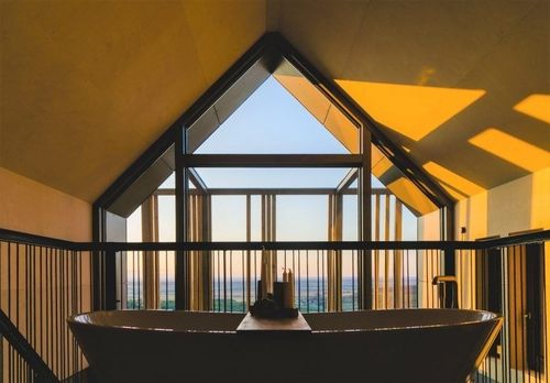 Eine Badewanne auf einem Balkon mit Blick auf den Sonnenuntergang. Ein großes dreieckiges Fenster sorgt für natürliches Licht.