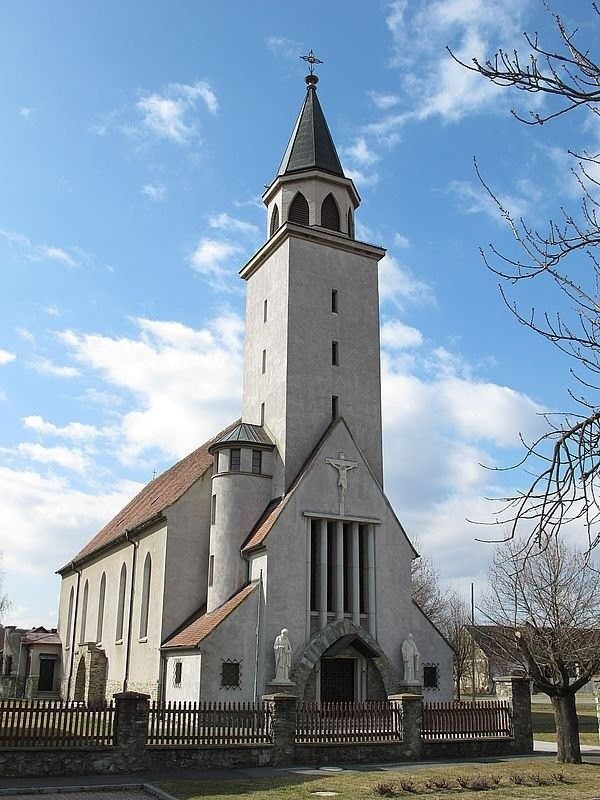 Eine weisse Kirche mit einem hohen Turm und einem Kruzifix an der Vorderseite. Sie hat ein rotes Dach und zwei Statuen auf beiden Seiten des Eingangs.