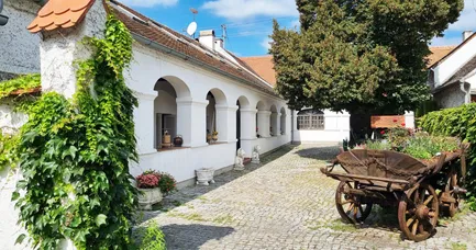 Ein traditionelles weißes Gebäude mit Bogenfenstern und einem braunen Dach, geschmückt mit Topfpflanzen und Löwenstatuen. Ein gepflasterter Weg führt zu einem hölzernen Wagen, mit einem großen Baum rechts.