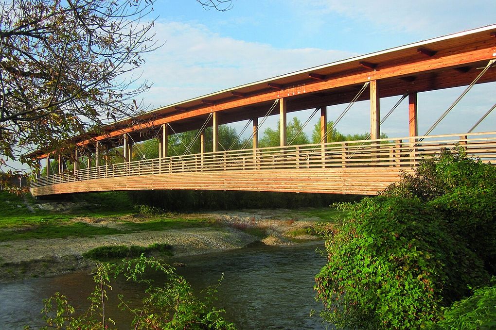  Abschnitt Bild: Eine lange Holzbrücke mit einem Metallgeländer, die einen ruhigen Fluss überspannt. Die Brücke wird von mehreren Metallträgern gestützt und ist von üppiger Grünfläche und einem klaren Himmel umgeben.