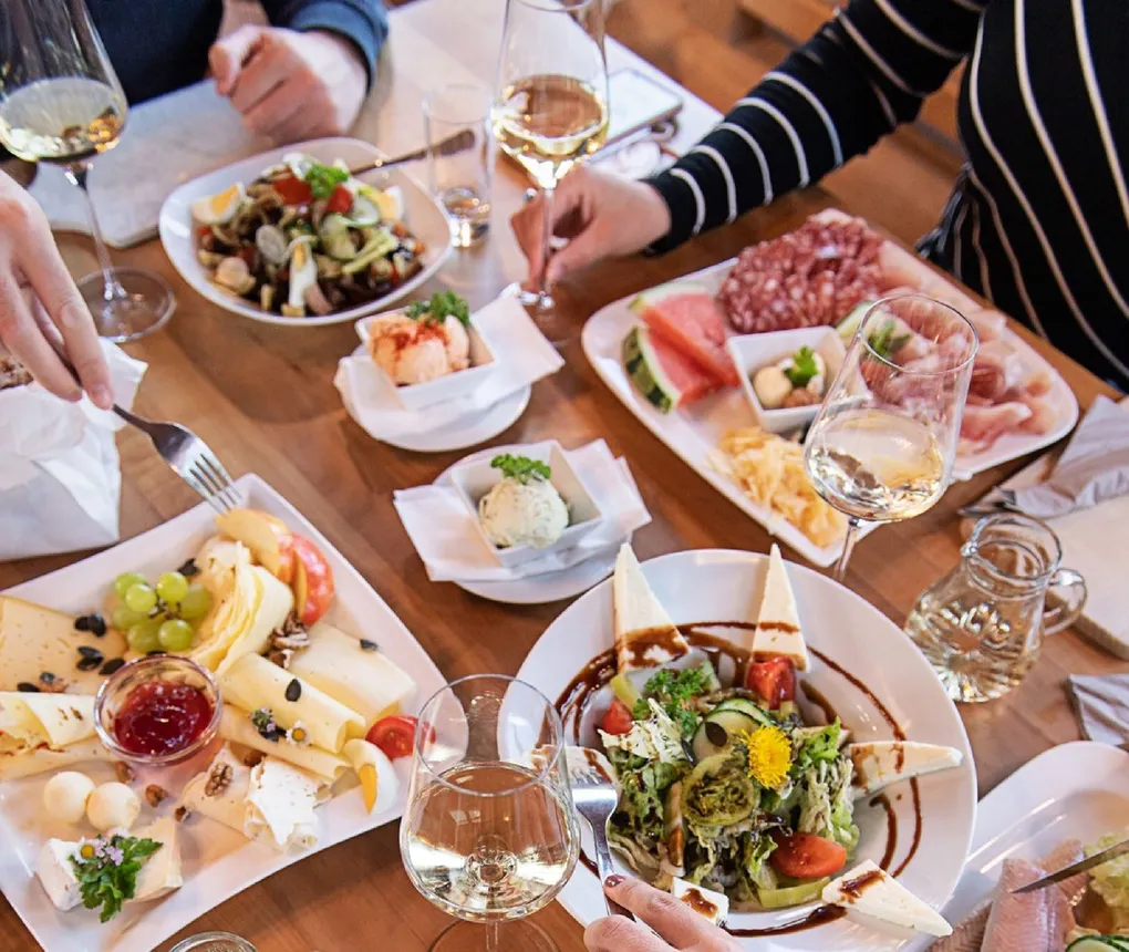 Abschnittsbild: Ein Holztisch mit verschiedenen Gerichten und Weingläsern. Eine Person hält ein Glas Wein und eine andere eine Gabel.