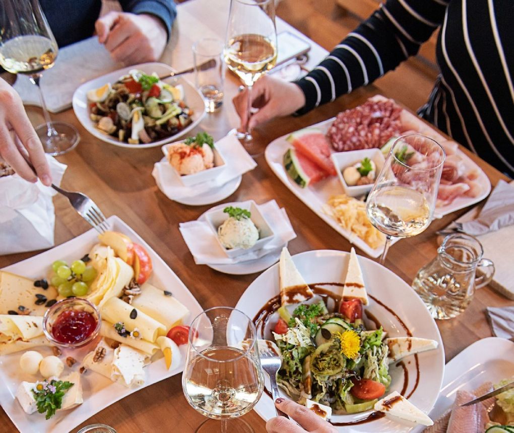  Abschnitt Bild: Ein Holztisch mit verschiedenen Gerichten und Weingläsern. Eine Person hält ein Glas Wein und eine andere eine Gabel.