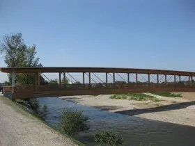 Eine lange Holzbrücke mit Metallrahmen überspannt einen Fluss unter blauem Himmel.