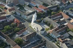 Luftaufnahme einer Stadt mit vielen Gebäuden, einige mit braunen Dächern, und einer weißen Kirche mit Turm in der Mitte.