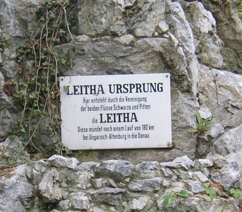 Ein Schild an einem felsigen Abhang mit der Aufschrift 'LeithA Ursprung' beschreibt, wie der Fluss Leitha durch die Vereinigung der Flüsse Schwarza und Pitten entsteht und nach 180 km bei Ungarisch-Altenburg in die Donau mündet.