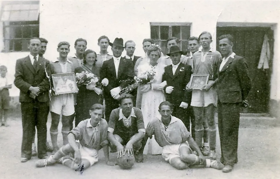 Eine Gruppe von Menschen in formeller Kleidung, wahrscheinlich ein Sportteam, posiert für ein Foto mit einem frisch verheirateten Paar in der Mitte. Der Mann und die Frau halten Blumen, während andere gerahmte Fotos und einen Fußball halten.