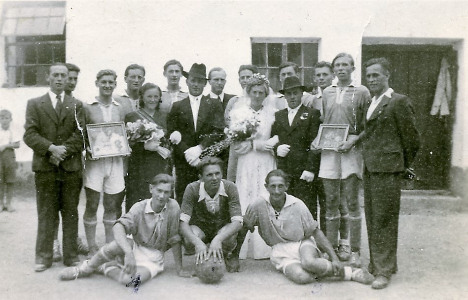 Eine Gruppe von Menschen in formeller Kleidung, wahrscheinlich ein Sportteam, posiert für ein Foto mit einem frisch verheirateten Paar in der Mitte. Der Mann und die Frau halten Blumen, während andere gerahmte Fotos und einen Fußball halten.