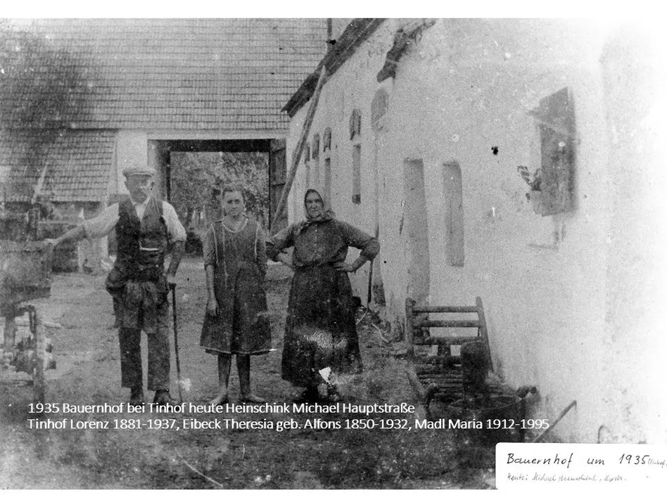 Schwarz-Weiß-Foto von drei Personen, die vor einem Bauernhaus stehen, mit dem Text 'Bauernhof bei Tinhof heute Heinschink Michael Hauptstraße' und 'Lorenz 1881-1937, Eibeck Theresia geb. Alfons 1850-1932, Madl Maria 1912-1995' unten.