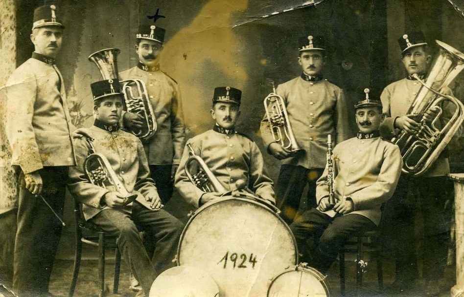 Eine Gruppe von Soldaten in Uniform, die Musikinstrumente halten. Zwei sitzend mit Trommeln, zwei stehend mit Trompeten und einer mit einem Tuba.