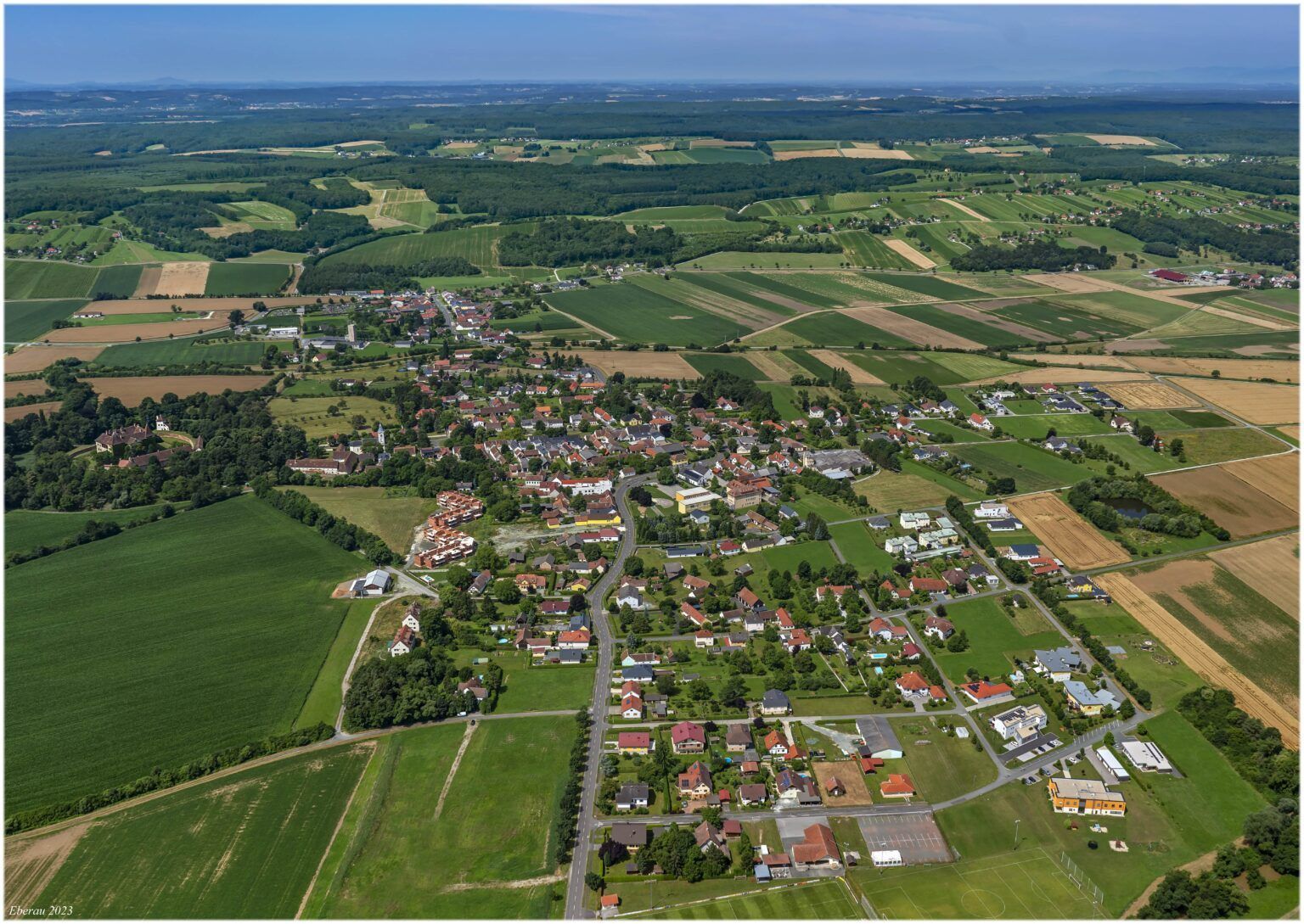 Luftaufnahme eines ländlichen Dorfes mit vielen Häusern, grünen Feldern und Bäumen, umgeben von einer weiten Landschaft.