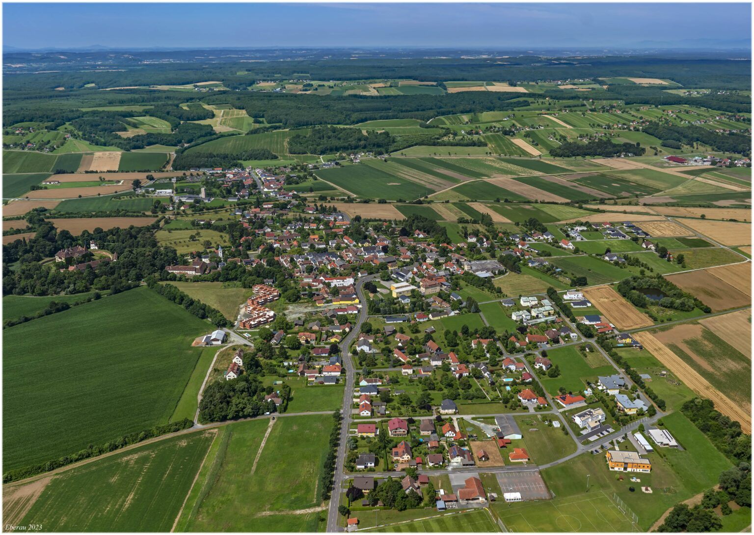 Luftaufnahme eines ländlichen Dorfes mit vielen Häusern, grünen Feldern und Bäumen, umgeben von einer weiten Landschaft.