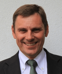 Ein Mann in einem schwarzen Anzug und einer grünen Krawatte lächelt in die Kamera. Er hat kurze braune Haare und steht vor einer hellen Wand.