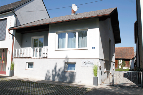 Ein weißes Haus mit braunem Dach hat einen Balkon mit weißen Geländern. An der Wand befindet sich ein Schild mit der Aufschrift Landhaus. Vor dem Haus stehen zwei Topfpflanzen. Das Haus hat einen Schornstein. Das Haus ist von einer Kiesauffahrt umgeben.