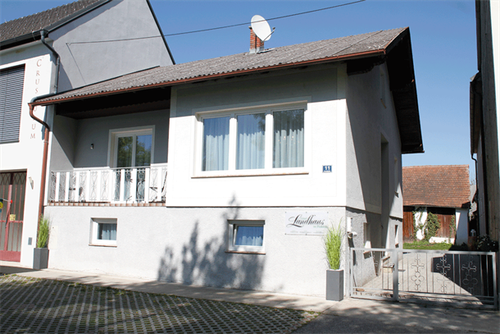 Ein weißes Haus mit braunem Dach hat einen Balkon mit weißen Geländern. An der Wand befindet sich ein Schild mit der Aufschrift Landhaus. Vor dem Haus stehen zwei Topfpflanzen. Das Haus hat einen Schornstein. Das Haus ist von einer Kiesauffahrt umgeben.