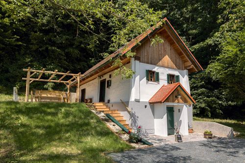 Ein weißes Haus mit Holzschindeln auf dem Dach befindet sich auf einem Grashügel. Das Haus hat einen hölzernen Eingang mit Treppenstufen. Eine kleine Veranda mit einer Bank befindet sich auf der linken Seite.