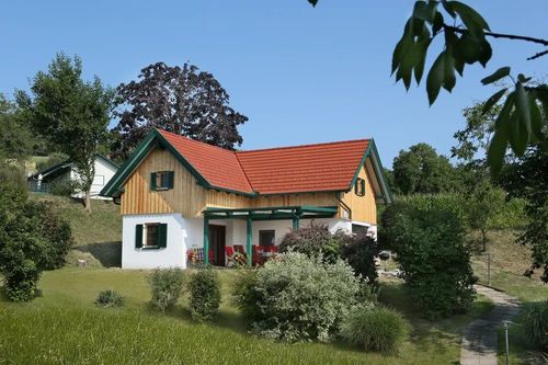 Ein holzverkleidetes Haus mit rotem Dach und grüner Einfassung steht in einem grünen Feld, umgeben von Bäumen und Büschen.